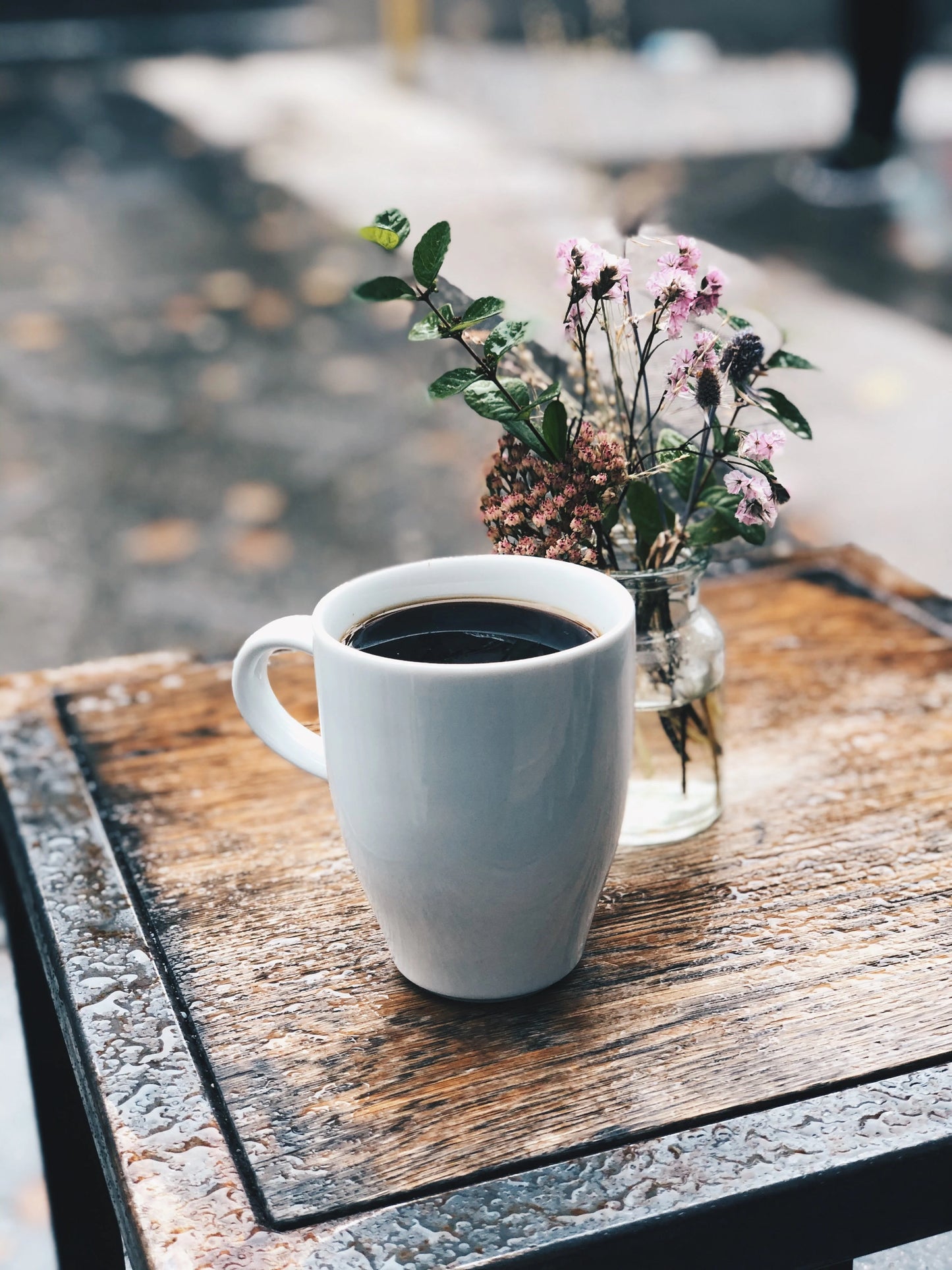 White mug with coffee and a small vase of flowers on a wooden table.